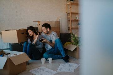 Wall Mural - Young couple is moving in to their new home. Young man is apologizing to his wife because he took phone call while they were drinking coffee on the floor of their new home.