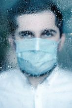 Blurred Image Of Young Man In Protective Mask Standing Behind The Window With Rain Drops.