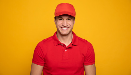 Wall Mural - Handsome young happy courier or delivery man in red uniform is posing isolated on yellow background.