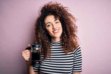 Young beautiful woman with curly hair and piercing doing coffee holding french coffeemaker with a happy face standing and smiling with a confident smile showing teeth