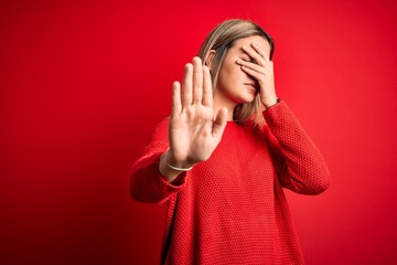 Poster - Young beautiful blonde woman wearing casual sweater over red isolated background covering eyes with hands and doing stop gesture with sad and fear expression. Embarrassed and negative concept.