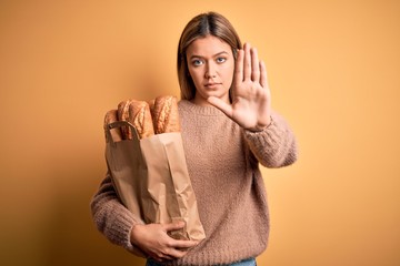 Sticker - Young beautiful woman holding paper bag with bread over isolated yellow background with open hand doing stop sign with serious and confident expression, defense gesture