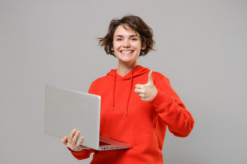 Smiling young brunette woman girl in casual red hoodie posing isolated on grey background studio portrait. People lifestyle concept. Mock up copy space. Working on laptop pc computer showing thumb up.