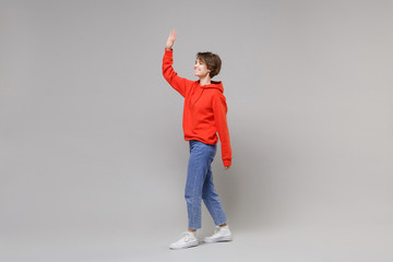 Wall Mural - Side view of joyful young brunette woman in casual red hoodie blue jeans posing isolated on grey background. People lifestyle concept. Mock up copy space. Waving greeting with hand as notices someone.