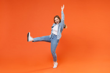 Cheerful young woman girl in casual denim clothes posing isolated on orange background studio portrait. People emotions lifestyle concept. Mock up copy space. Spreading hands, rising leg, dancing.