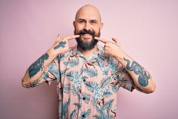 Poster - Handsome bald man with beard and tattoo wearing casual floral shirt over pink background smiling cheerful showing and pointing with fingers teeth and mouth. Dental health concept.