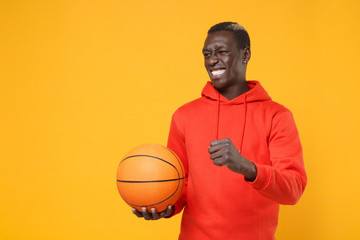 Irritated young african american man guy basketball player in red streetwear hoodie posing isolated on yellow background. Sport leisure lifestyle concept. Playing basketball hold ball clenching fist.