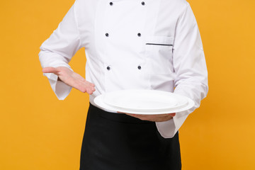 Cropped image of young male chef cook or baker man in white uniform shirt posing isolated on yellow background studio portrait. Cooking food concept. Mock up copy space. Pointing hand on empty plates.