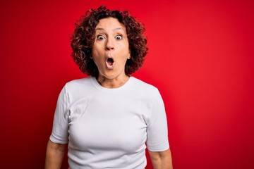 Poster - Middle age beautiful curly hair woman wearing casual t-shirt over isolated red background afraid and shocked with surprise expression, fear and excited face.