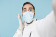 © ViDi Studio - Close up of shocked male doctor man in medical gown face mask gloves isolated on blue background. Epidemic pandemic coronavirus 2019-ncov sars covid-19 flu virus concept. Doing selfie on mobile phone.