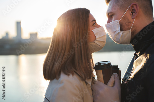 Photographie Young couple, a man and a woman in medical masks and gloves, drink coffee from disposable cups on the street and look at each other against the background of the river and the city