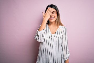 Wall Mural - Young beautiful woman wearing casual striped t-shirt and glasses over pink background covering one eye with hand, confident smile on face and surprise emotion.