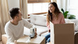 © fizkes - Happy mixed race woman talking with smiling husband while packing home stuff into carton boxes for moving in new house. Joyful family couple preparing belongings for delivery, relocation service.