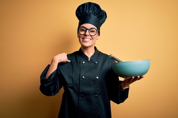 Poster - Young beautiful brunette chef woman wearing cooker uniform and hat holding bowl and whisk with surprise face pointing finger to himself