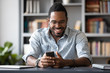 Overjoyed millennial African American man in glasses sit at desk laugh texting or typing on cellphone, happy biracial male in spectacles have fun watch funny video on smartphone gadget indoors