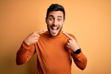Wall Mural - Young handsome man with beard wearing casual sweater standing over yellow background smiling cheerful showing and pointing with fingers teeth and mouth. Dental health concept.