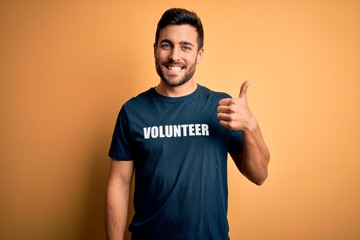 Wall Mural - Young handsome man with beard volunteering wearing t-shirt with volunteer message happy with big smile doing ok sign, thumb up with fingers, excellent sign