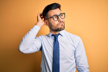 Young handsome businessman wearing tie and glasses standing over yellow background confuse and wondering about question. Uncertain with doubt, thinking with hand on head. Pensive concept.