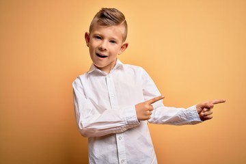 Young little caucasian kid with blue eyes wearing elegant white shirt over yellow background smiling and looking at the camera pointing with two hands and fingers to the side.