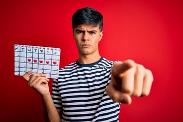 Young handsome man holding calendar with hearts over isolated red background pointing with finger to the camera and to you, hand sign, positive and confident gesture from the front