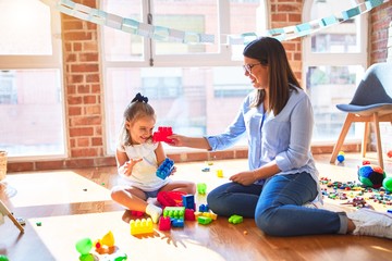 Poster - Caucasian girl kid playing and learning at playschool with female teacher. Mother and daughter at playroom around toys playing with bulding blocks