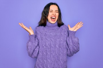 Young beautiful woman wearing casual turtleneck sweater standing over purple background celebrating mad and crazy for success with arms raised and closed eyes screaming excited. Winner concept