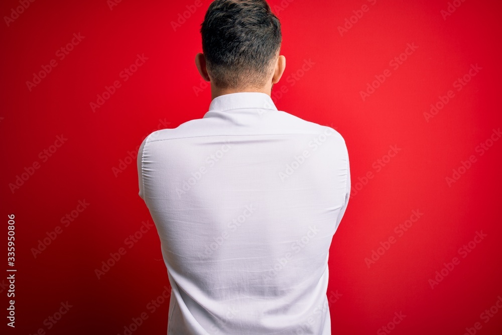 Young business man with blue eyes wearing elegant shirt standing over red isolated background standing backwards looking away with crossed arms