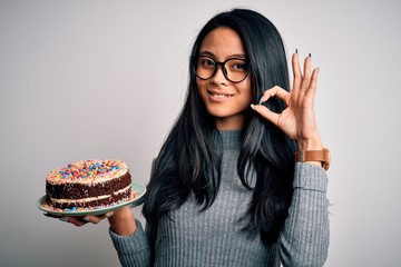 Young beautiful chinese woman holding birthday cake standing over isolated white background doing ok sign with fingers, excellent symbol