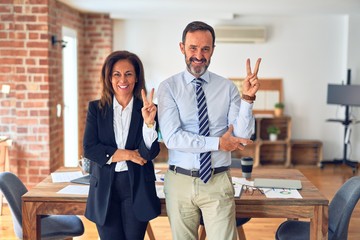 Two middle age business workers standing working together in a meeting at the office smiling with happy face winking at the camera doing victory sign. Number two.