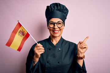 Middle age patriotic chef woman wearing cooker uniform and hat holding spain spanish flag very happy pointing with hand and finger to the side