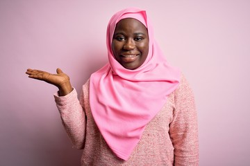 Canvas Print - Young african american plus size woman wearing muslim hijab over isolated pink background smiling cheerful presenting and pointing with palm of hand looking at the camera.