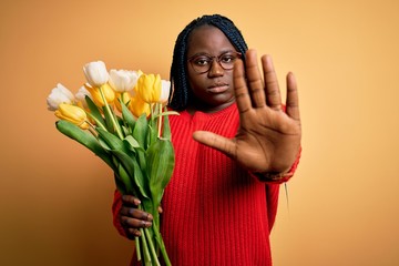 Poster - Young african american plus size woman with braids holding bouquet of yellow tulips flower doing stop sing with palm of the hand. Warning expression with negative and serious gesture on the face.