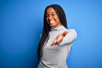 Sticker - Young african american woman standing wearing casual turtleneck over blue isolated background smiling cheerful offering palm hand giving assistance and acceptance.