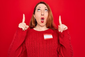 Poster - Young beautiful redhead woman wearing sticker presentation with hello my name message amazed and surprised looking up and pointing with fingers and raised arms.