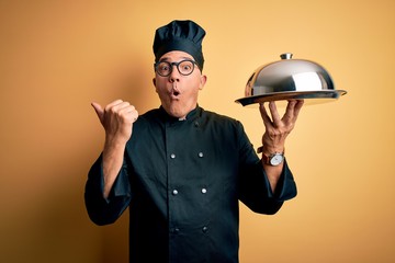 Poster - Middle age handsome grey-haired waiter man wearing cooker uniform and hat holding tray Surprised pointing with hand finger to the side, open mouth amazed expression.