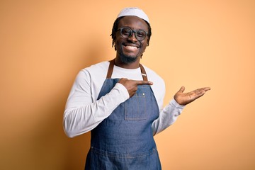 Sticker - Young african american cooker man wearing apron and over isolated yellow background amazed and smiling to the camera while presenting with hand and pointing with finger.