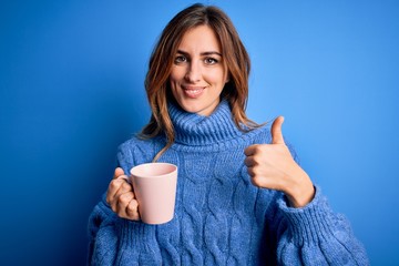 Young beautiful brunette woman drinking cup of coffe over isolated blue background happy with big smile doing ok sign, thumb up with fingers, excellent sign