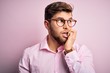 © Krakenimages.com - Young handsome blond man with beard and blue eyes wearing pink shirt and glasses looking stressed and nervous with hands on mouth biting nails. Anxiety problem.