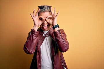 Young handsome modern man wearing fashion leather jacket and sunglasses over yellow background doing ok gesture like binoculars sticking tongue out, eyes looking through fingers. Crazy expression.