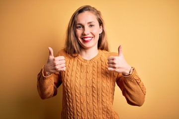 Young beautiful blonde woman wearing casual sweater standing over yellow background success sign doing positive gesture with hand, thumbs up smiling and happy. Cheerful expression and winner gesture.