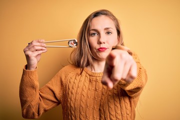 Wall Mural - Young beautiful blonde woman eating sushi using chopsticks over isolated yellow background pointing with finger to the camera and to you, hand sign, positive and confident gesture from the front