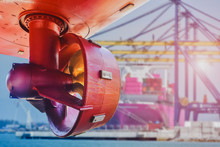 Red Propeller In Rope Guard With Rudder Under Ship Repairing In Shipyard Near Port And Crane Lifting Loading Cargo Container In Port Background.