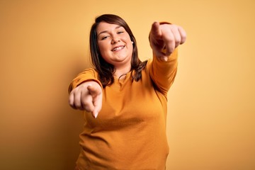 Beautiful brunette plus size woman wearing casual sweater over isolated yellow background pointing to you and the camera with fingers, smiling positive and cheerful