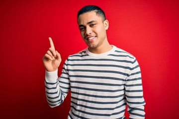 Young brazilian man wearing casual striped t-shirt standing over isolated red background pointing finger up with successful idea. Exited and happy. Number one.