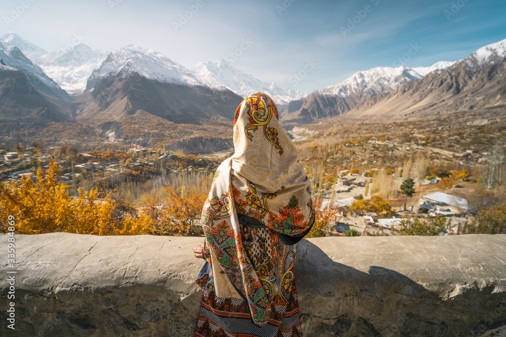 A woman wearing traditional dress sitting on wall and looking at Hunza valley in autumn season, Gilgit Baltistan in Pakistan - obrazy, fototapety, plakaty 