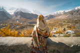 Fototapeta Krajobraz - A woman wearing traditional dress sitting on wall and looking at Hunza valley in autumn season, Gilgit Baltistan in Pakistan