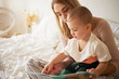 © shurkin_son - Indoor image of concentrated patient young mother sitting on white bed with little child on lap, holding book, teaching her son alphabet while social distancing at home because of quarantine