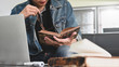 © Prathankarnpap - Close-up man holding and reading a book while sitting in front a computer laptop at the leather couch over comfortable living room as background.