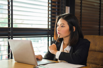  young Asian woman in formal suit sitting n front of laptop and holding a pencil learning her chin while thinking when looking outside window