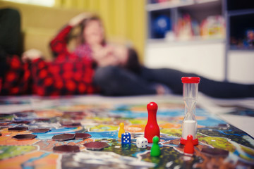 Wall Mural - Board game concept- friends spend time together. Board game field, figures, dice, coins and sandglass. Two people play holding cards on blurred background